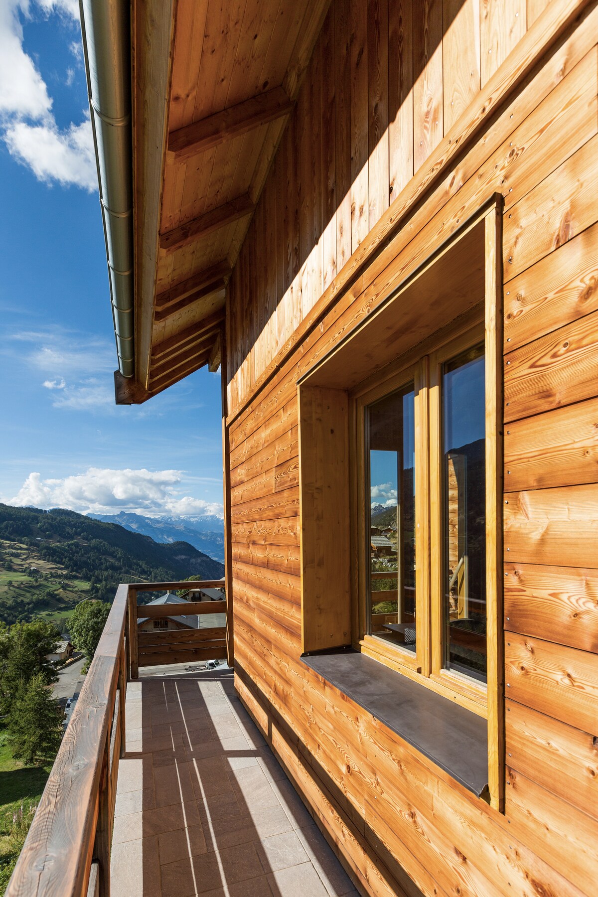 A wooden balcony is framed by natural wood paneling, offering views of the mountainous landscape. Large windows provide ample light and showcase the outdoor scenery. The sky features occasional clouds, enhancing the serene atmosphere of the location.