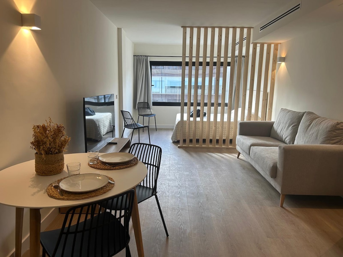 A modern living space features a cozy sofa and a dining table set for two. Divided by a wooden screen, the sleeping area includes a spacious bed with light bedding. Natural light enters through a large window, contributing to a bright and open atmosphere.