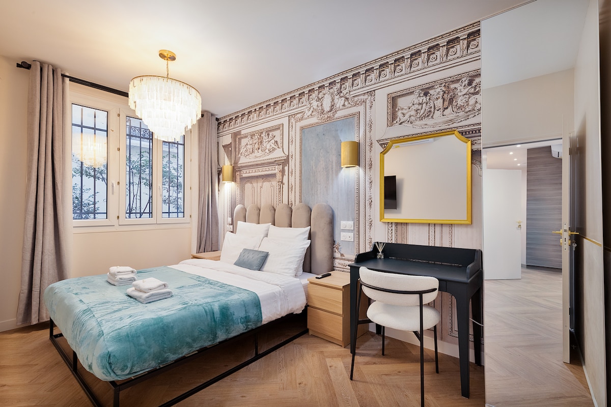 A modern bedroom features a spacious bed dressed in soft turquoise linens. A chandelier hangs above, complemented by golden wall sconces. A small desk with a mirror is positioned against the wall, while large windows allow natural light to fill the room, framed by light curtains.