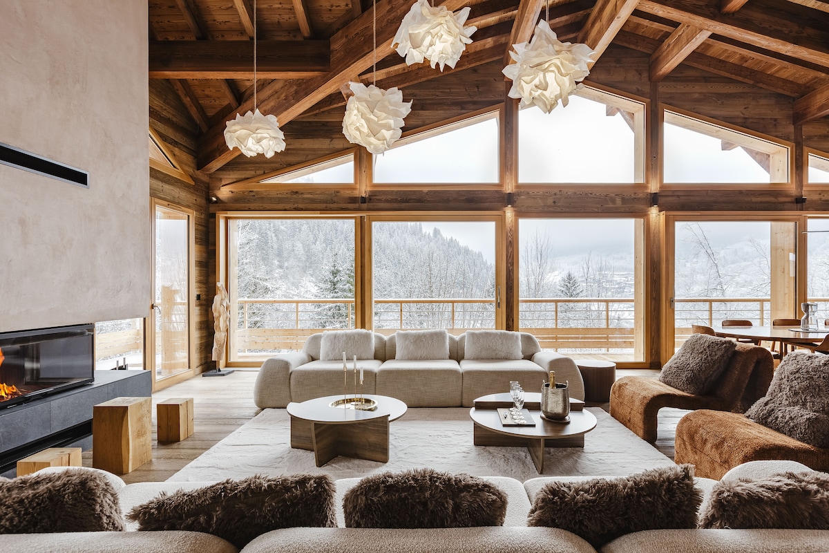 A spacious living area features large windows that frame a snowy landscape, allowing natural light to fill the room. The rustic wood beams complement cozy furnishings, including a light-colored sectional sofa and a warm-toned accent chair. A modern fireplace adds a touch of comfort to the atmosphere.