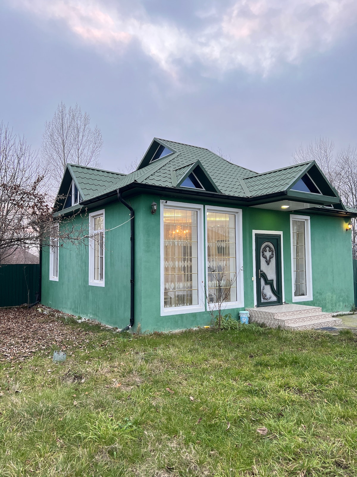 The exterior of the house features a unique green facade with white windows and a decorative front door. A well-maintained grassy area surrounds the property, and large windows allow natural light to brighten the interior.