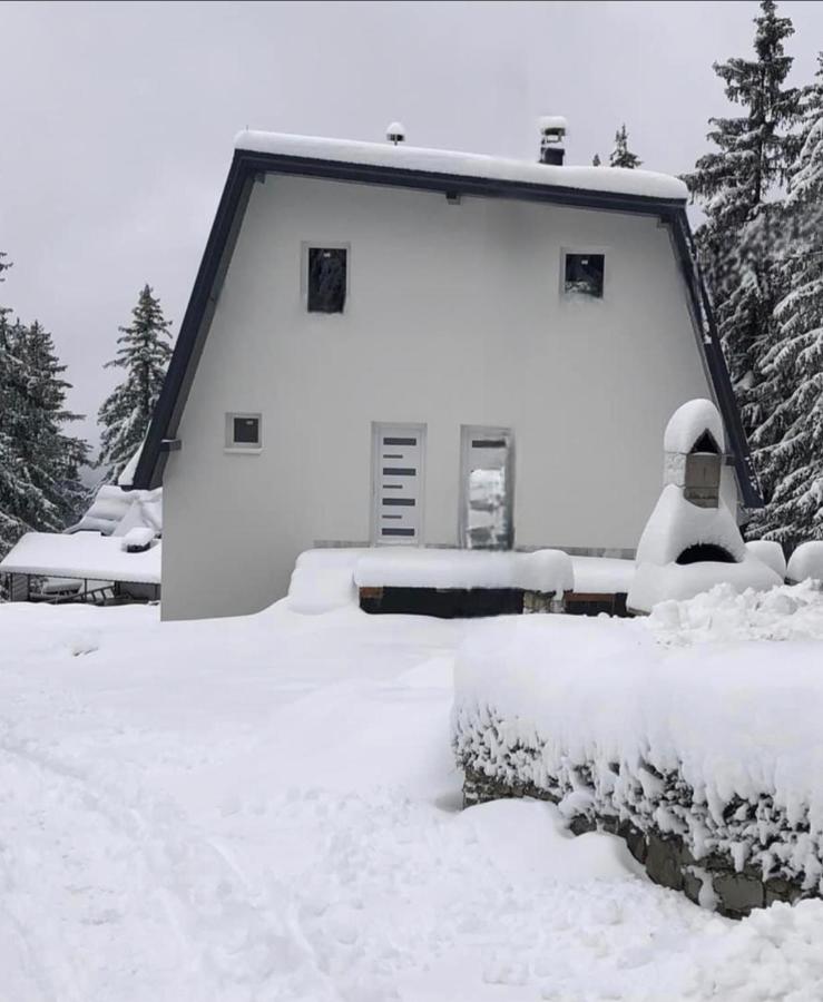A modern building is surrounded by snow-covered terrain, with a sloped roof and several windows visible. The entrance features a distinctive door design, while snow blankets the ground and nearby pathways, creating a serene winter landscape.