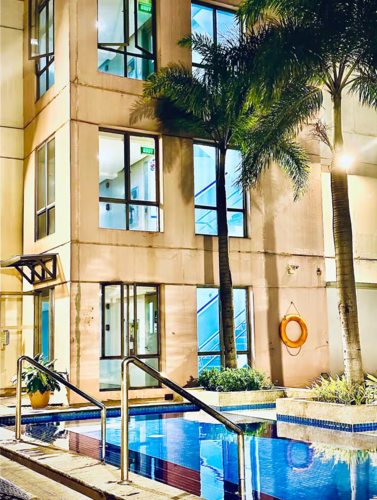 A well-lit pool area is bordered by a modern building featuring large glass windows. Lush palm trees provide greenery, while a life buoy is mounted on the wall nearby. The pool reflects the soft lighting from the surrounding area, offering a serene atmosphere.