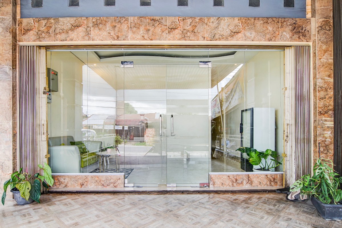 A glass entrance showcases a modern, welcoming lobby space. Indoor plants provide a touch of greenery, while comfortable seating is visible through the transparent doors. The flooring features a subtle geometric pattern, adding to the clean and spacious atmosphere.