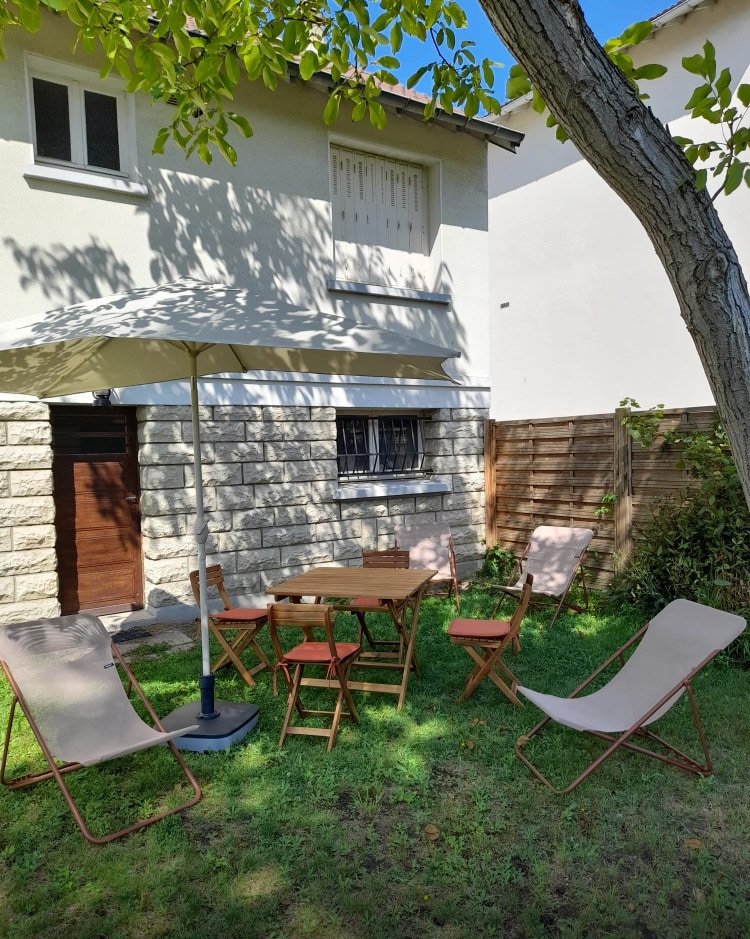 A shaded outdoor seating area is set up in the garden, featuring a wooden table and four folding chairs. An umbrella provides coverage from the sun, while green grass surrounds the seating arrangement. Nearby, a tree offers additional shade and greenery.