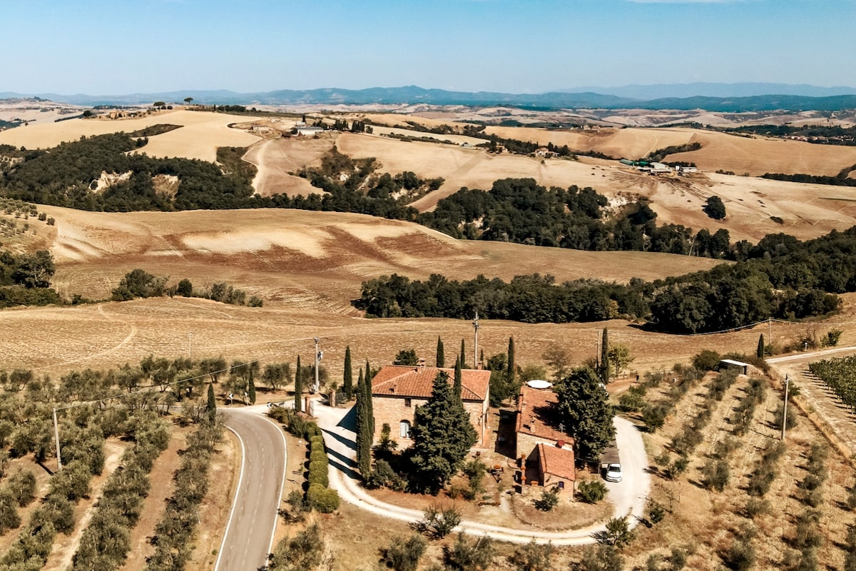 The image captures a scenic aerial view of the countryside, featuring rolling hills and cultivated fields. A rustic property surrounded by cypress trees is visible, with a winding road leading to it. Olive groves and vineyards enhance the landscape’s natural beauty.