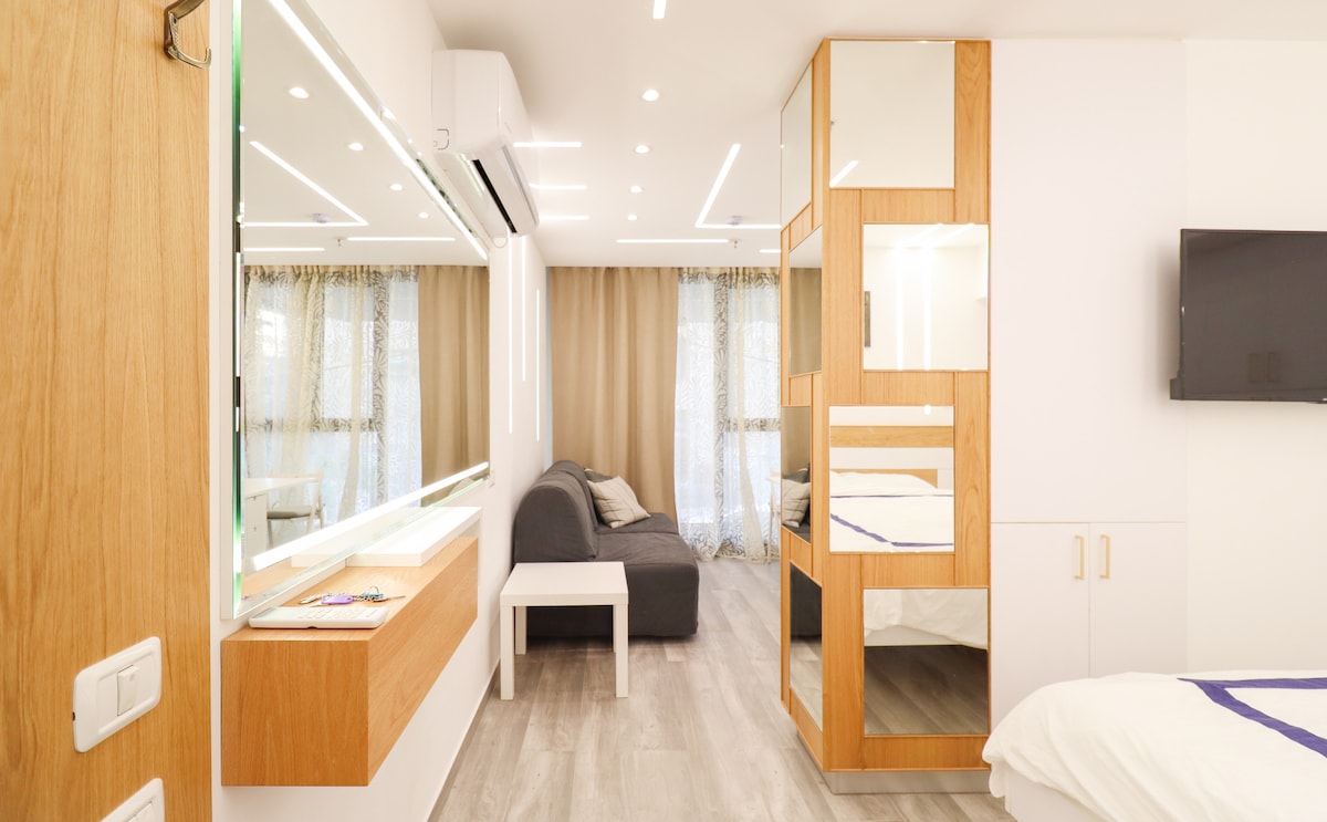 A modern interior showcases a spacious room with light wood accents. A comfortable gray sofa is positioned next to a sleek white table. Abundant natural light filters through sheer curtains, highlighting the reflective surfaces and clean lines of the design.
