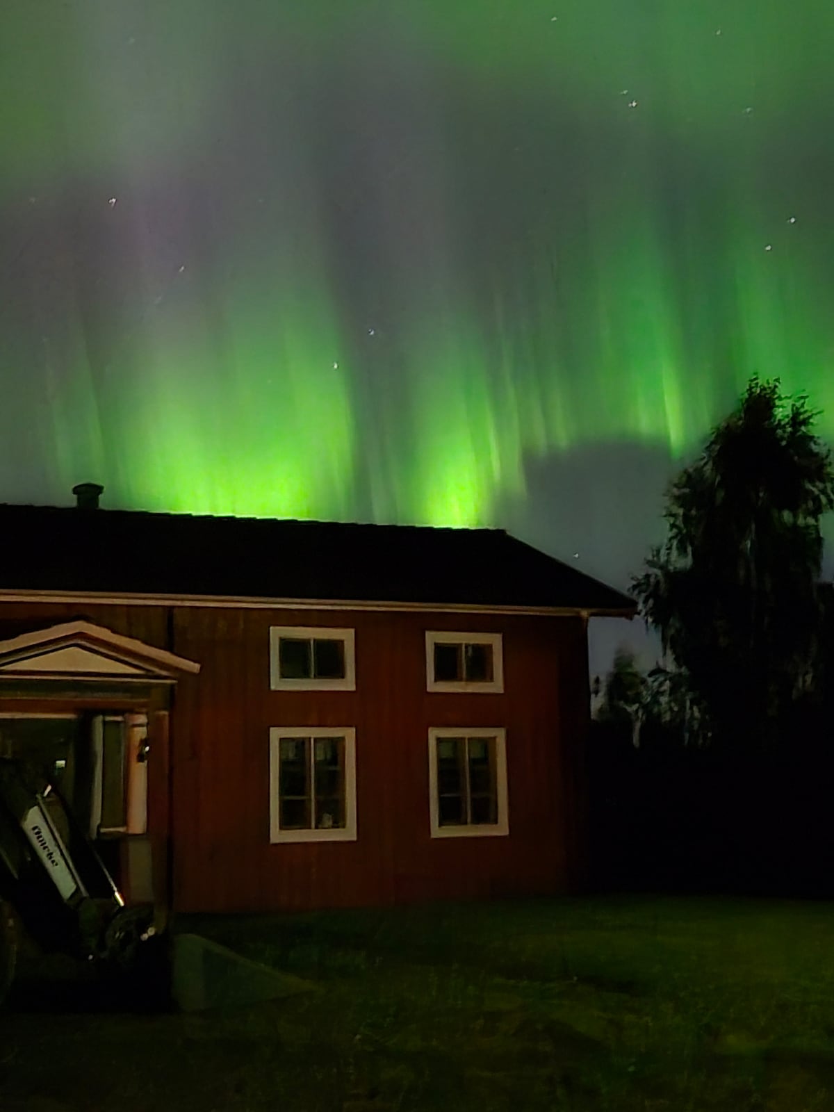 The night sky is illuminated by vibrant green auroras, casting a serene glow over the surrounding landscape. A rustic building with a dark roof and several windows is positioned on the left, while tall trees frame the right side of the image, enhancing the tranquil setting.