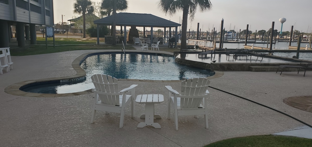 A relaxing outdoor area is highlighted by a shimmering pool surrounded by lounge chairs. Two white Adirondack chairs are positioned at the edge, facing a quaint table. A covered gazebo is visible nearby, complementing the marina views in the background.