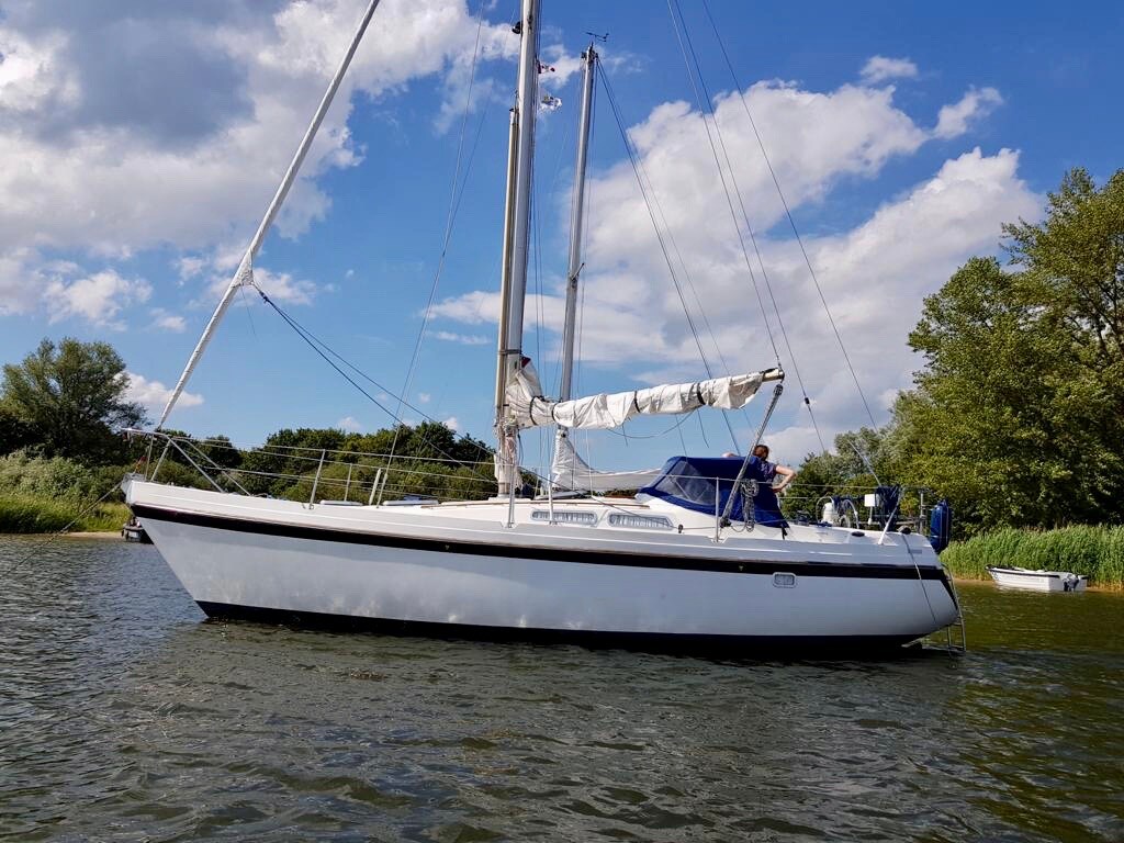 A sleek white sailboat is anchored on calm waters, surrounded by lush greenery and scattered clouds in a blue sky. The boat features a canopy over the cockpit and a tall mast with sails that are furled.