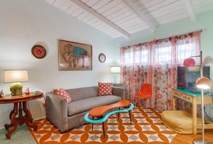 A cozy living area is highlighted by vaulted ceilings and a cheerful color palette. A gray sofa is accented with colorful pillows, accompanied by mid-century modern furnishings. Natural light filters through patterned curtains, illuminating the unique patterned rug beneath.