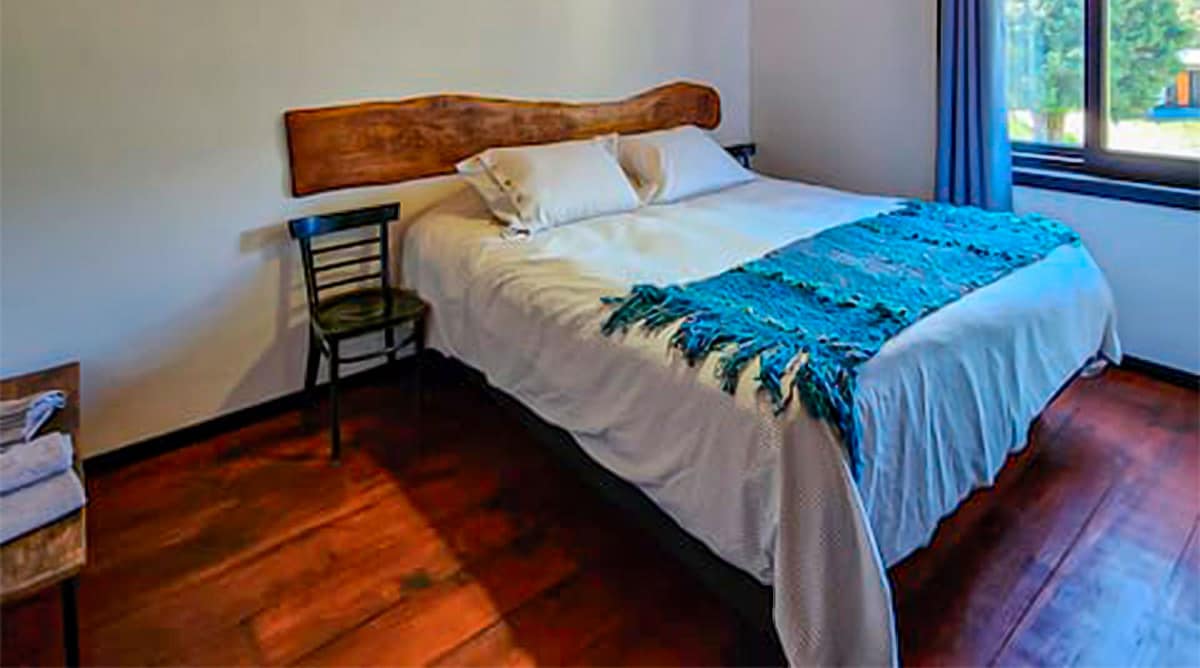 A cozy sleeping area is highlighted by a spacious bed with a wooden headboard. Soft pillows rest atop the bed, and a colorful blanket is draped over the edge. Natural light enters through the window, and a chair and small side table are positioned nearby.