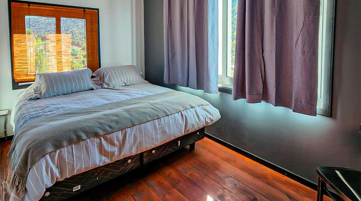 A spacious bedroom features a large bed with a light-colored blanket. Natural light filters through two windows, one adorned with bamboo shades and the other with dark curtains. The wooden floor complements the warm tones of the room.