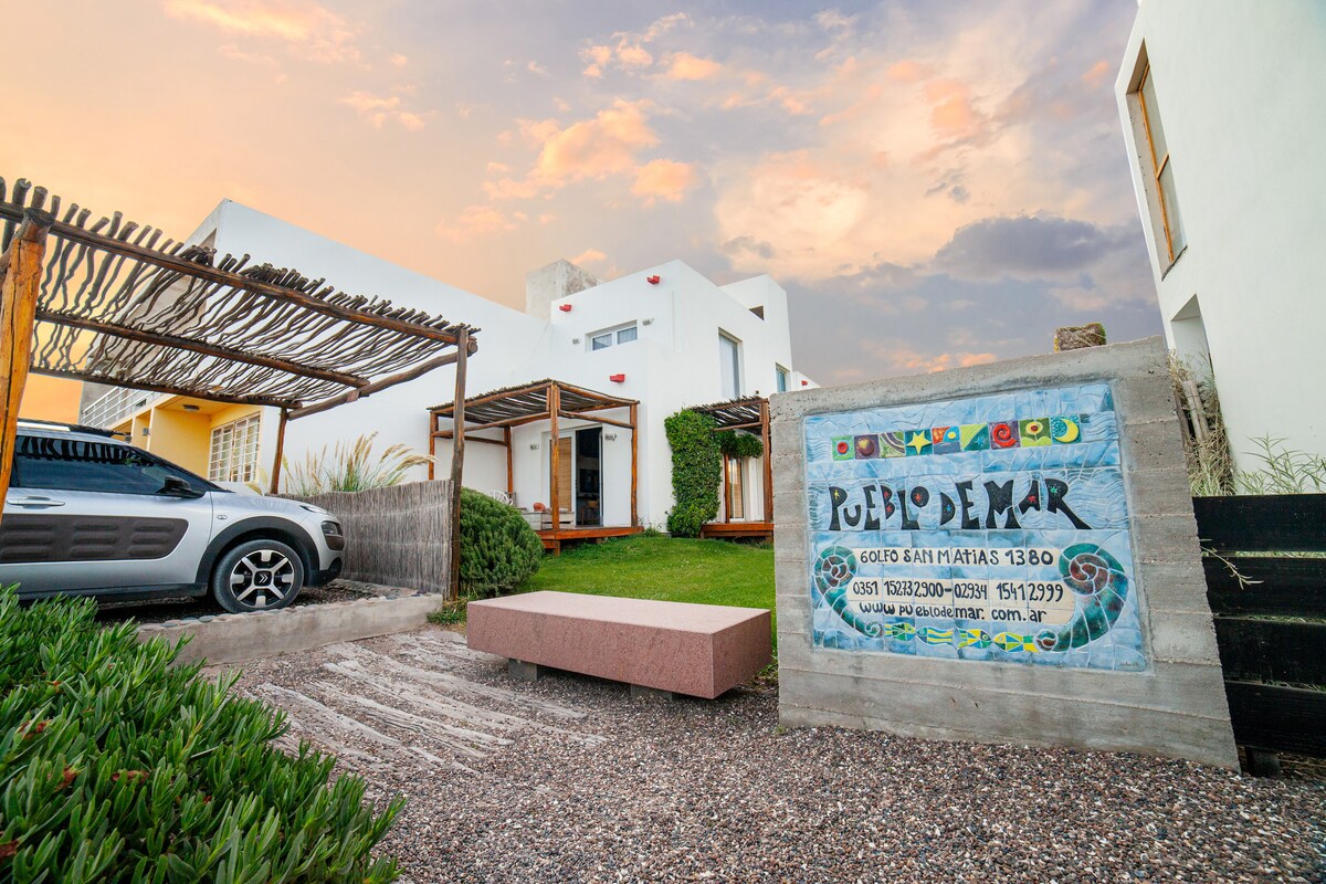 A glimpse of the Pueblo de Mar complex is presented, featuring well-maintained gardens and a gravel pathway leading to the entrance. A sign displays the name of the complex, while a parked vehicle offers a sense of accessibility. The backdrop showcases the colorful sky at sunset.