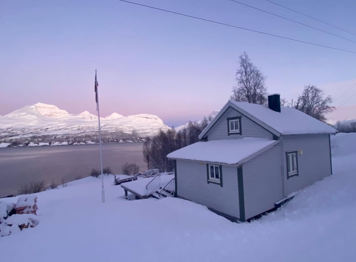 A charming house is set against a backdrop of snow-covered mountains and a serene body of water. The surrounding landscape is blanketed in fresh snow, while a flagpole stands in front of the house. Soft hues of dawn light illuminate the scene.