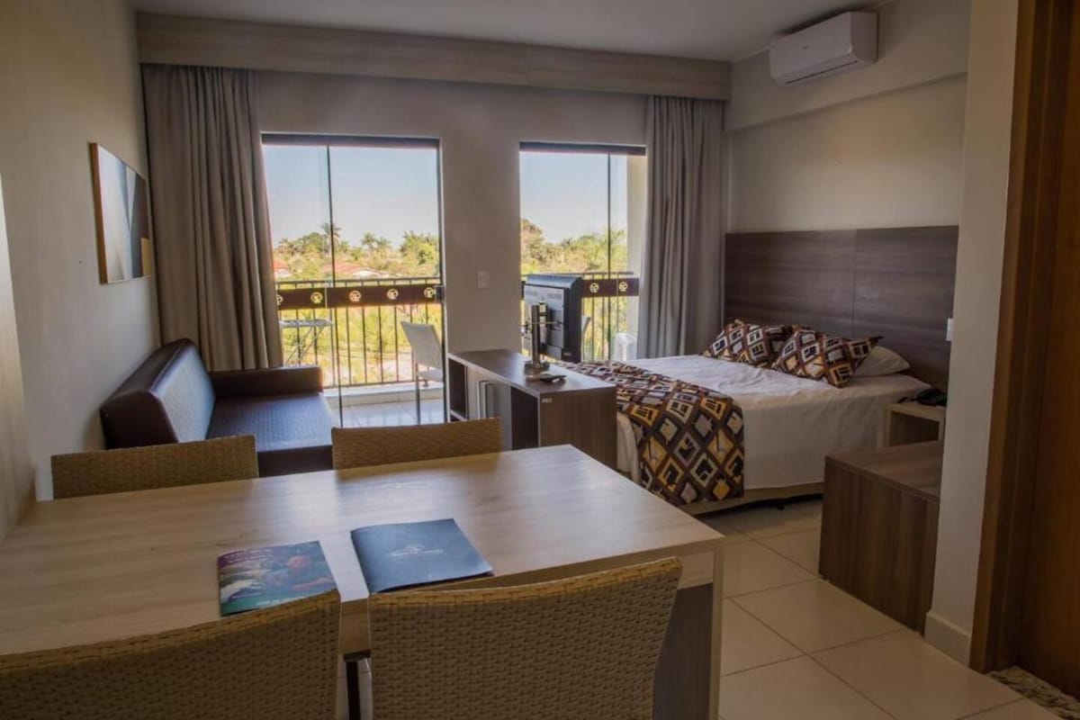 A well-lit room features a spacious bed with patterned bedding, accompanied by a sofa and a dining table. Large windows provide natural light and views of the outdoors. A refrigerator is placed near the bed, while air conditioning is visible above for comfort.