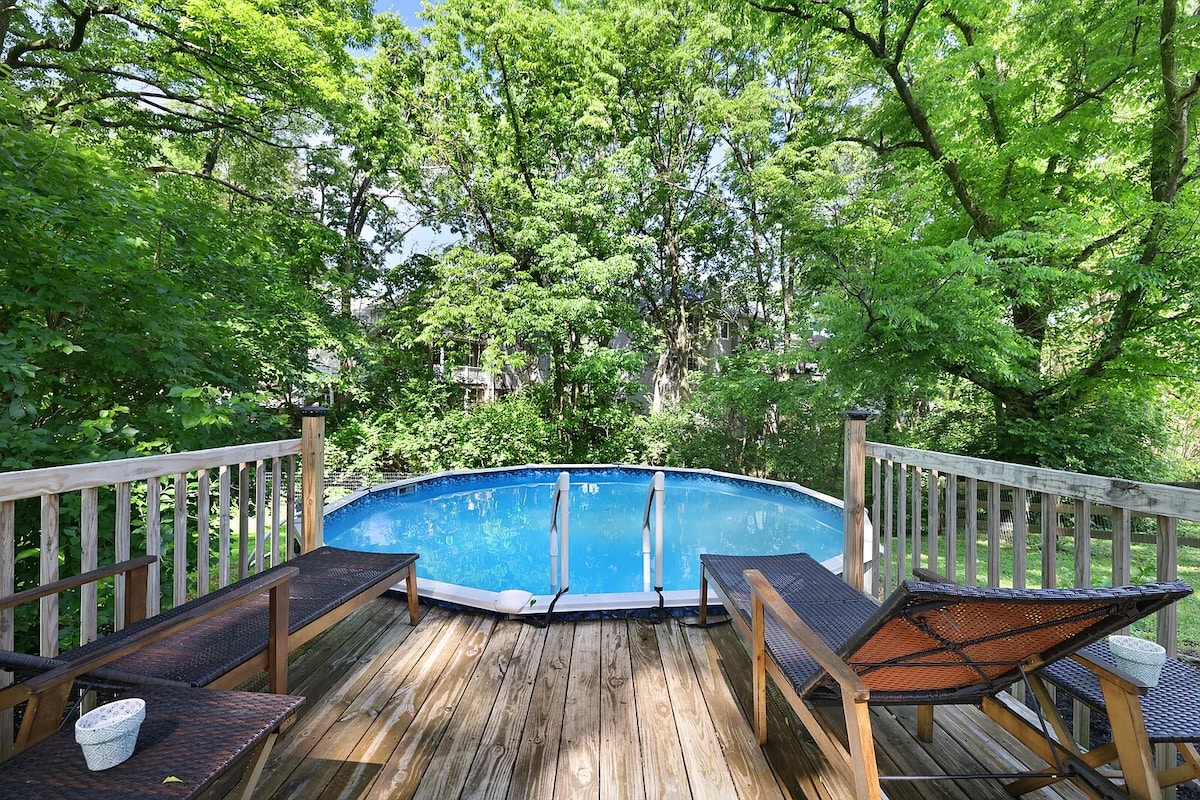 A wooden deck surrounds a refreshing above-ground pool, inviting relaxation. Two lounge chairs are positioned nearby for comfort, with lush green trees providing a natural backdrop and shade. The clear blue water of the pool contrasts beautifully with the surrounding greenery.