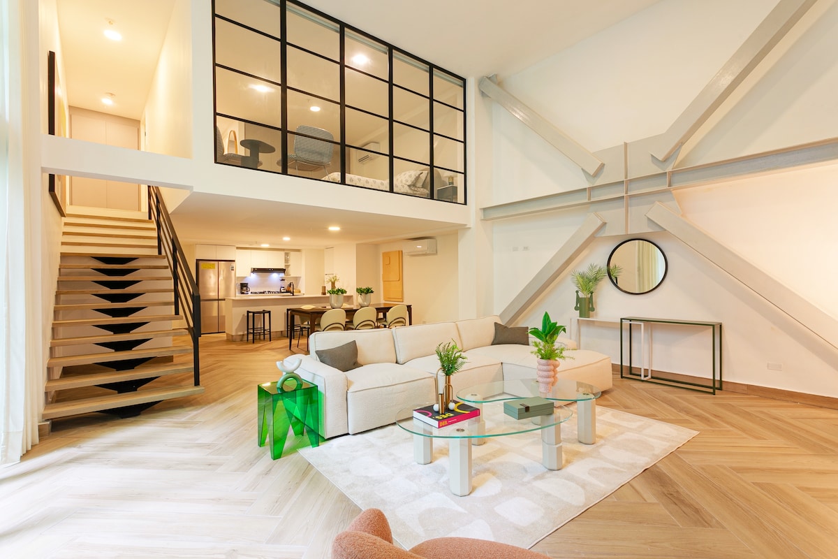 The inviting living area features a modern sectional sofa set against a large glass wall, offering a view of the upper floor. A glass coffee table rests on a textured area rug. The space is brightened by natural light reflecting off light wood flooring and a stylish mirror on the wall.