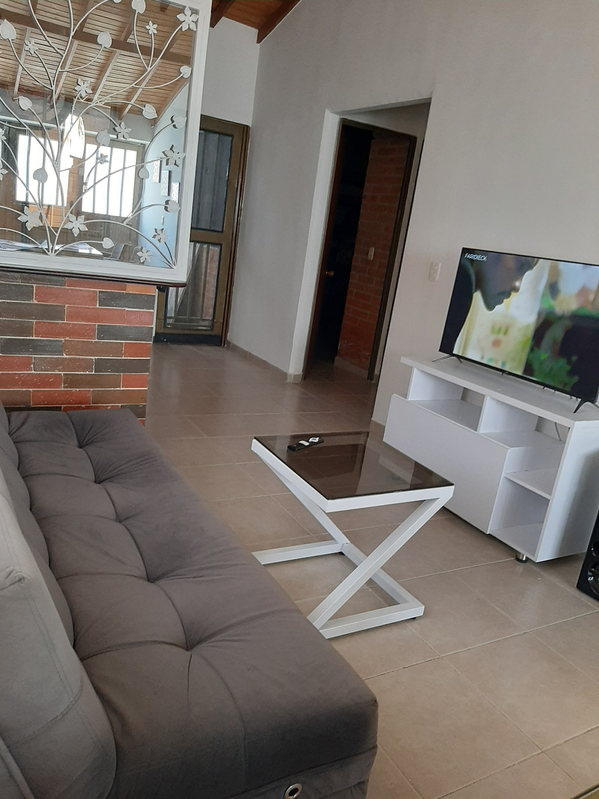 A modern living area features a gray sofa positioned near a sleek white coffee table. A large Smart TV is on an entertainment unit against the wall. Natural light filters through large windows, illuminating the tiled floor and creating a relaxed environment.