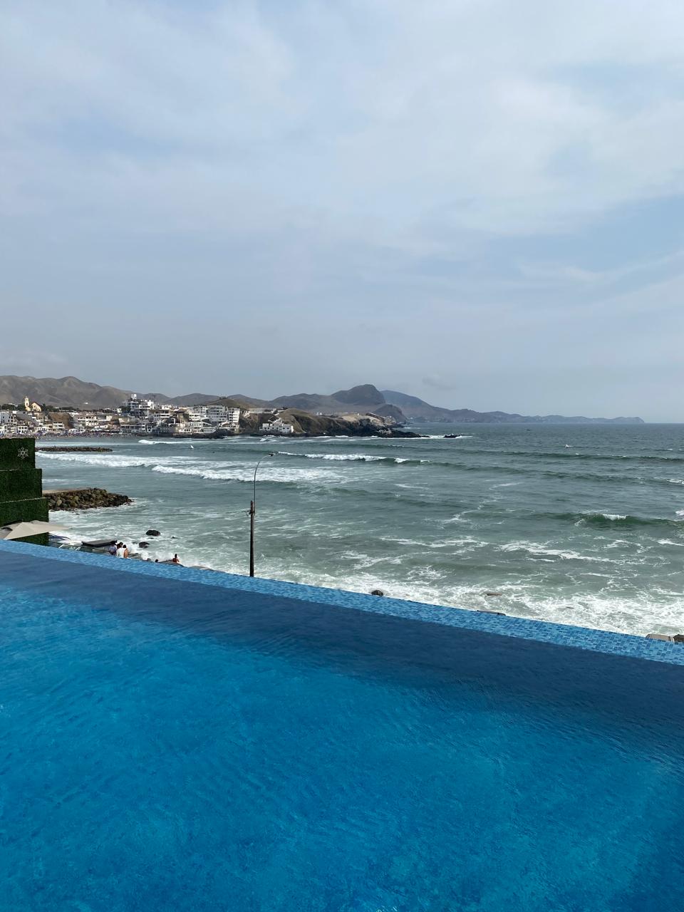 An expansive infinity pool appears to blend seamlessly with the ocean, offering views of rolling waves and a distant shoreline. The surrounding landscape includes hills and a quaint coastal town, creating a serene backdrop for relaxation.