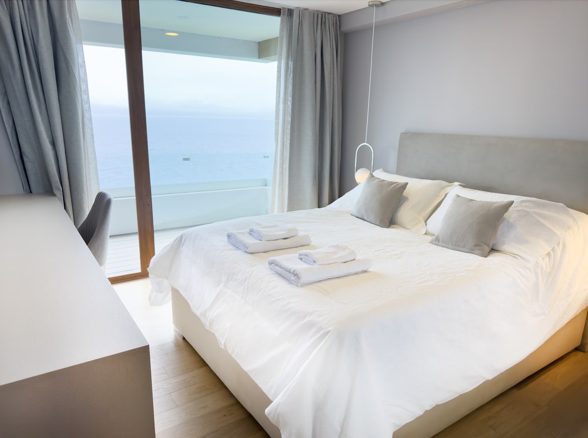 A bright and airy bedroom features a spacious bed with neatly arranged towels. Soft gray curtains frame a large glass door, revealing a picturesque view of the ocean. A minimalist desk adds a functional element to the modern, serene design.