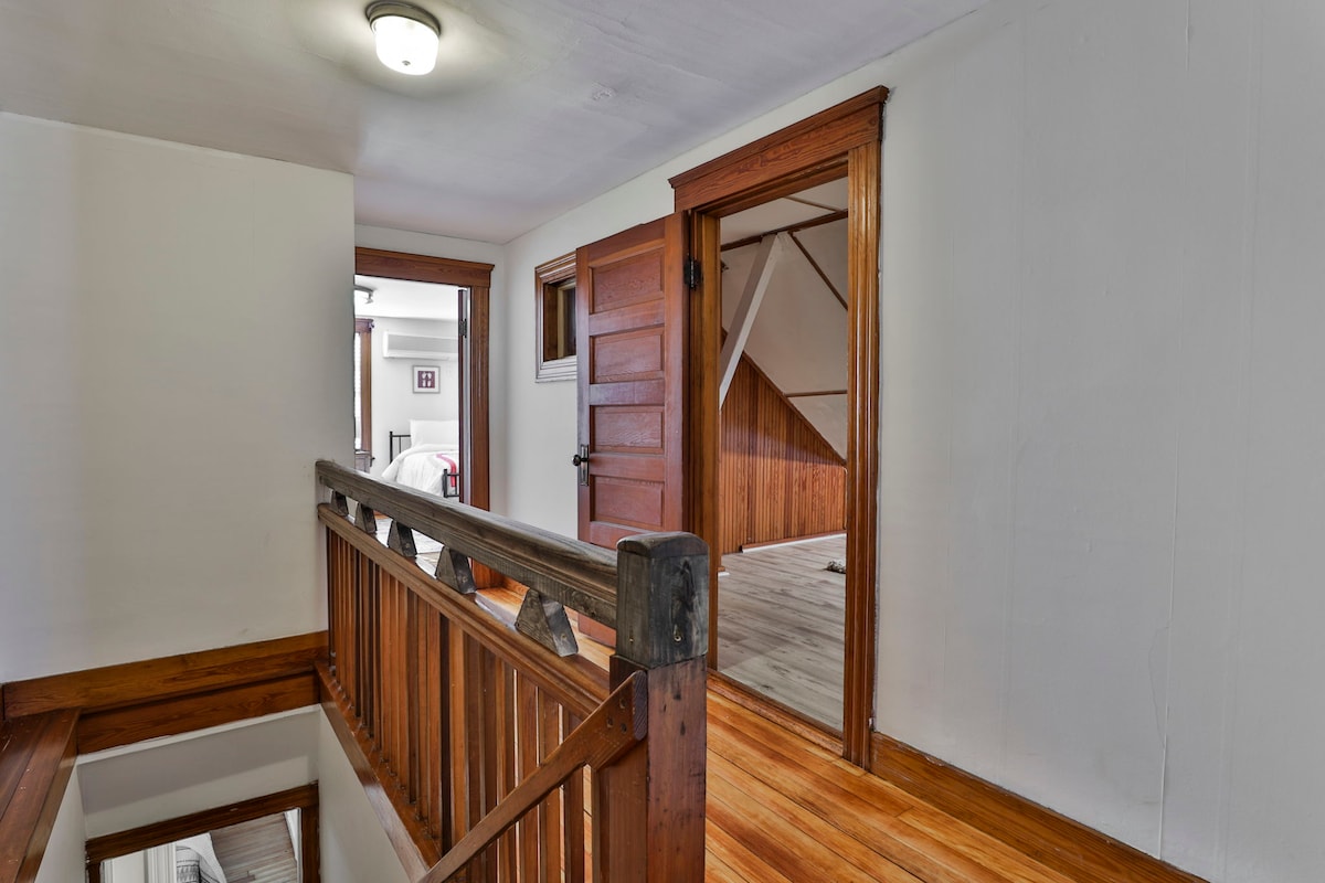 A second-floor hallway features polished wooden floors and natural light streaming through an opening. Rustic wood trim outlines the doorways and railing, leading to an entryway with a sloped ceiling. The space offers access to additional rooms, enhancing the sense of openness.