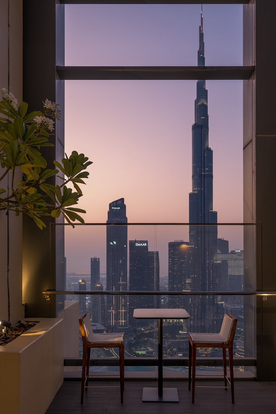 A cozy dining setup is positioned by a large window, offering an unobstructed view of the Burj Khalifa against a sunset sky. Two chairs are paired with a small table, while potted greenery adds a touch of nature to the scene.