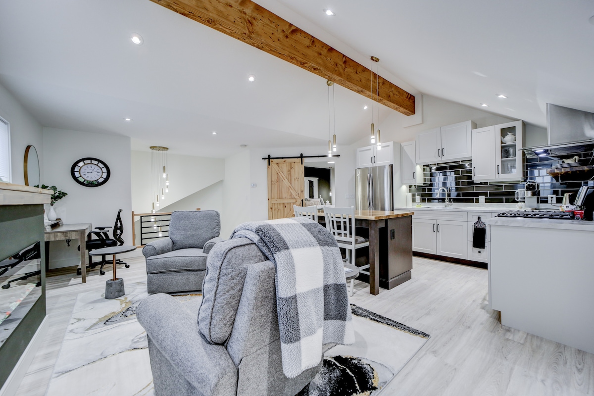 A spacious living area features a modern kitchen equipped with stainless steel appliances and a large island. Cozy seating is provided by a pair of gray chairs, with natural light coming through multiple windows. A decorative area rug enhances the inviting atmosphere.