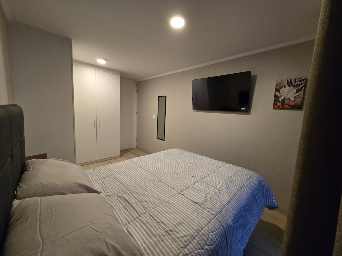 A comfortable bedroom is equipped with a spacious bed covered in light-colored linens. A wall-mounted television is positioned across from the bed. A wardrobe is visible in the corner, along with a small artwork displayed on an adjacent wall, contributing to a functional layout.