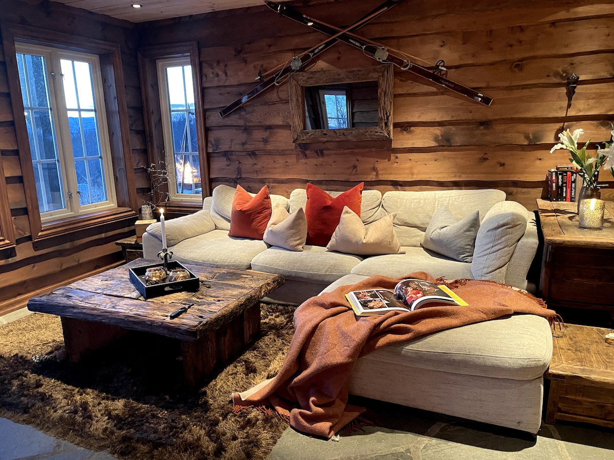A spacious seating area is highlighted by a light-colored sectional sofa adorned with several decorative pillows. A rustic wooden coffee table sits in front, with an open magazine displayed. Soft lighting from the windows illuminates the wooden walls, creating a cozy atmosphere.