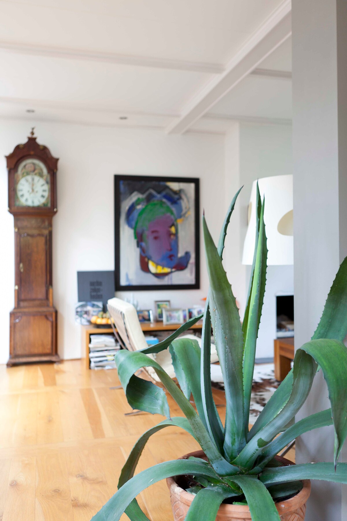 A living area showcases a tall indoor plant, adding a touch of nature to the space. A vintage clock stands against the wall, complemented by contemporary art and a cozy seating arrangement. Light hardwood flooring and soft illumination enhance the inviting atmosphere.