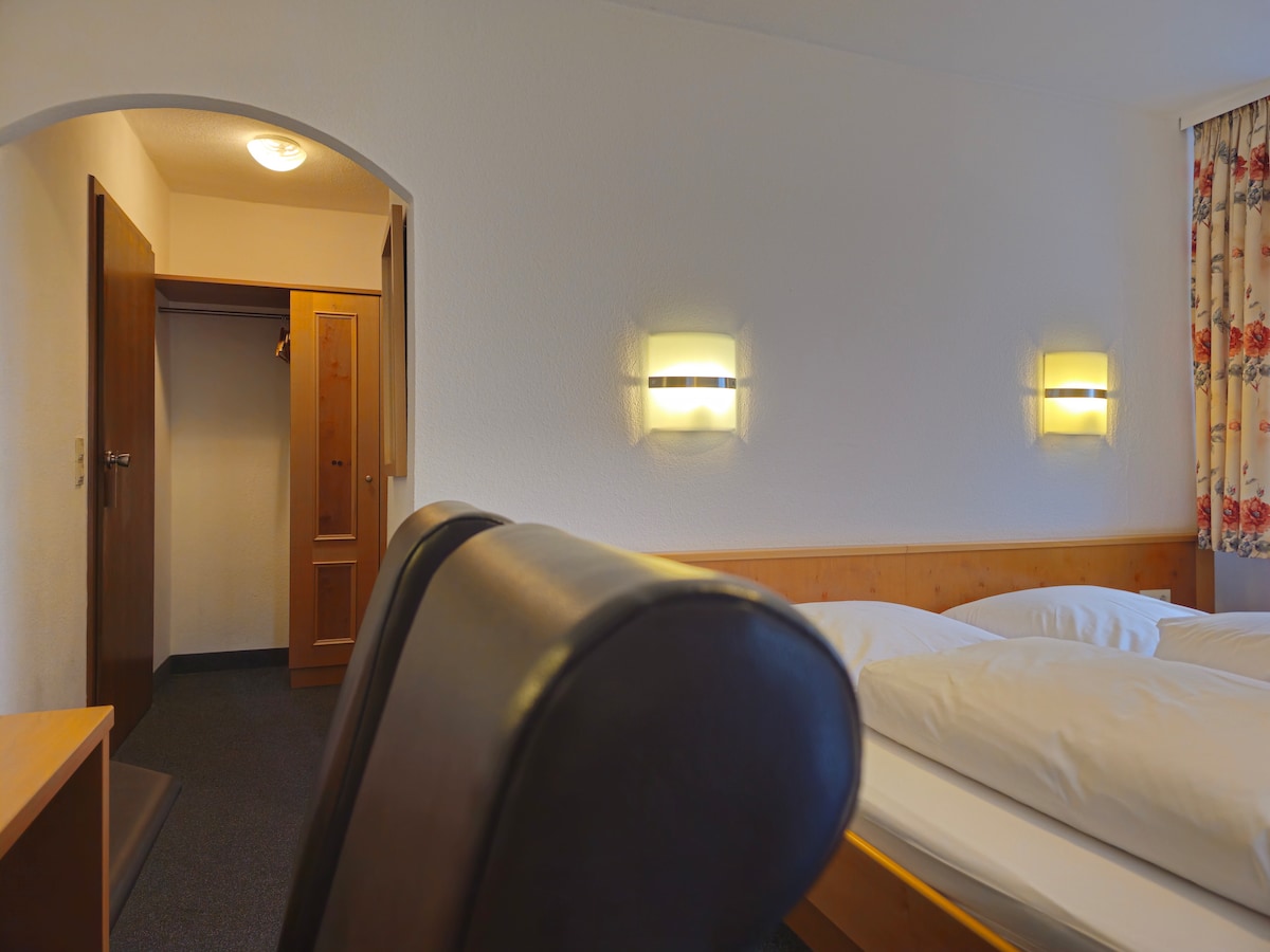 A double room is presented with a spacious bed and wooden accents. A closet is visible to the left, alongside a simple writing desk and chair. Soft lighting from wall fixtures creates a calm atmosphere, while a patterned curtain adorns the window.