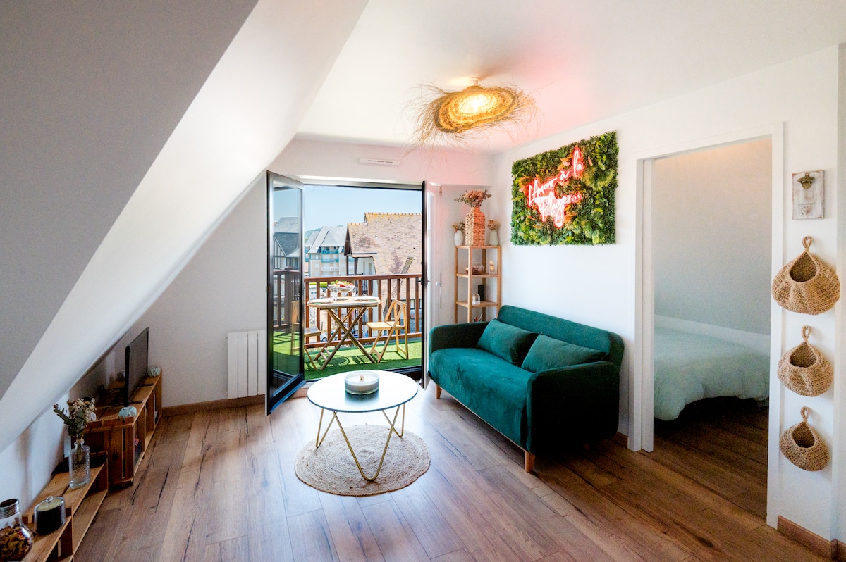 A bright living area features a green sofa and a circular coffee table placed on a wooden floor. Large glass doors open to a balcony with outdoor seating. Wall decor adds color, while natural light floods the space through the vaulted ceiling.