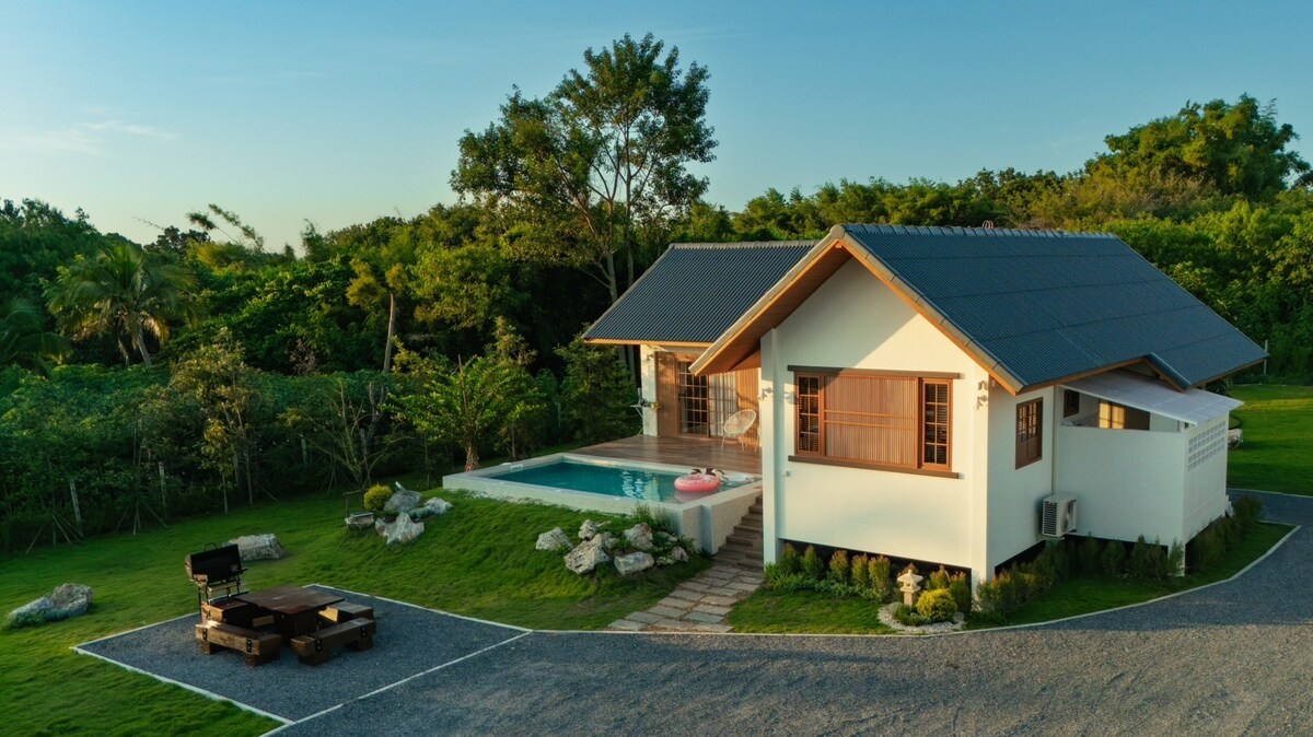 The villa features a modern design with a sloped roof and large windows. An outdoor saltwater pool is visible, surrounded by lush greenery. A wooden deck lounge area is located to the left, offering a relaxing space amidst the well-maintained garden.