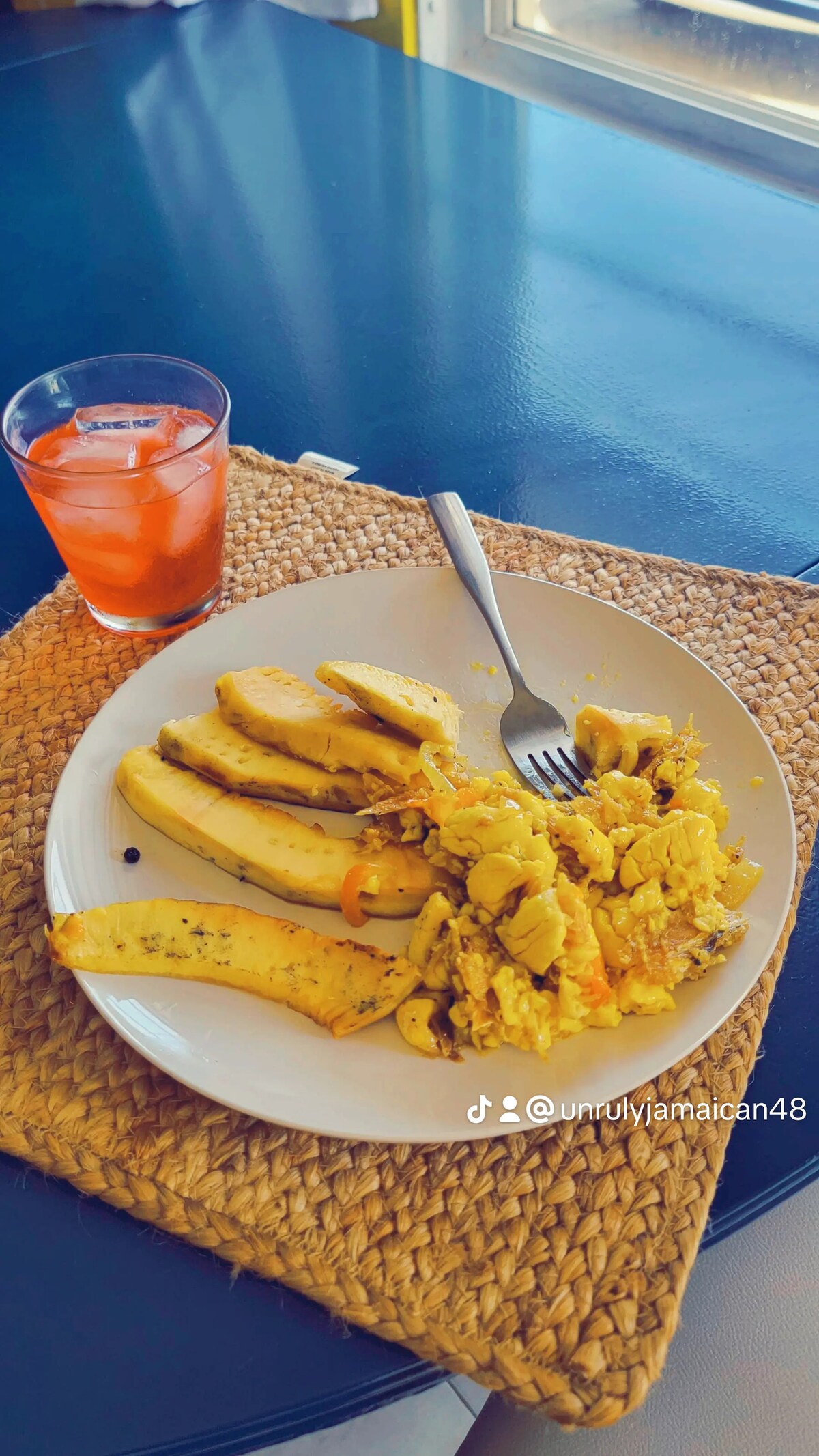 A plate holds a serving of scrambled eggs with vegetables alongside several pieces of cooked plantains. A clear glass with a light beverage sits nearby on a woven placemat, with a modern table surface visible underneath.