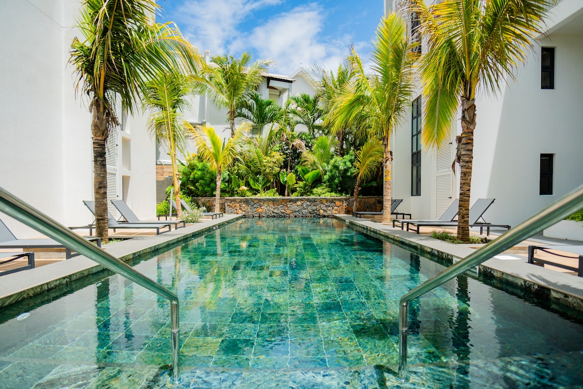 A modern swimming pool is framed by palm trees, reflecting clear blue skies above. Comfortable lounge chairs are positioned alongside the water, offering a peaceful retreat. Lush greenery surrounds the area, providing a tranquil setting for relaxation.