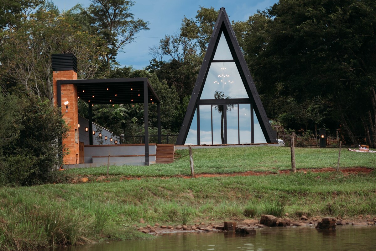 The exterior of a modern triangular chalet is displayed, featuring large glass panels and a contrasting brick chimney. Surrounded by lush greenery, it overlooks a tranquil lake, reflecting the natural landscape and offering an inviting outdoor seating area with lights strung across.