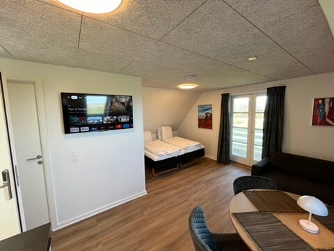 A modern room is displayed, featuring a flat-screen TV mounted on the wall opposite a bed setup with two single mattresses. Natural light enters through the large window, and stylish art pieces adorn the walls. A cozy sitting area includes a round table and chairs.