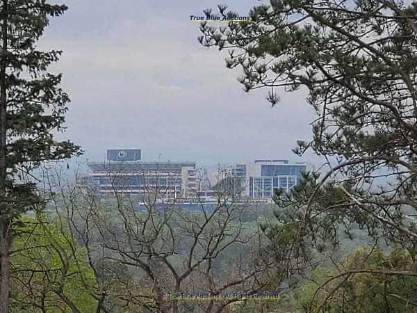 A distant view of a large stadium is framed by surrounding trees. The stadium is partially obscured, suggesting its size and significance. A soft haze blankets the scene, adding a subtle atmosphere to the landscape.