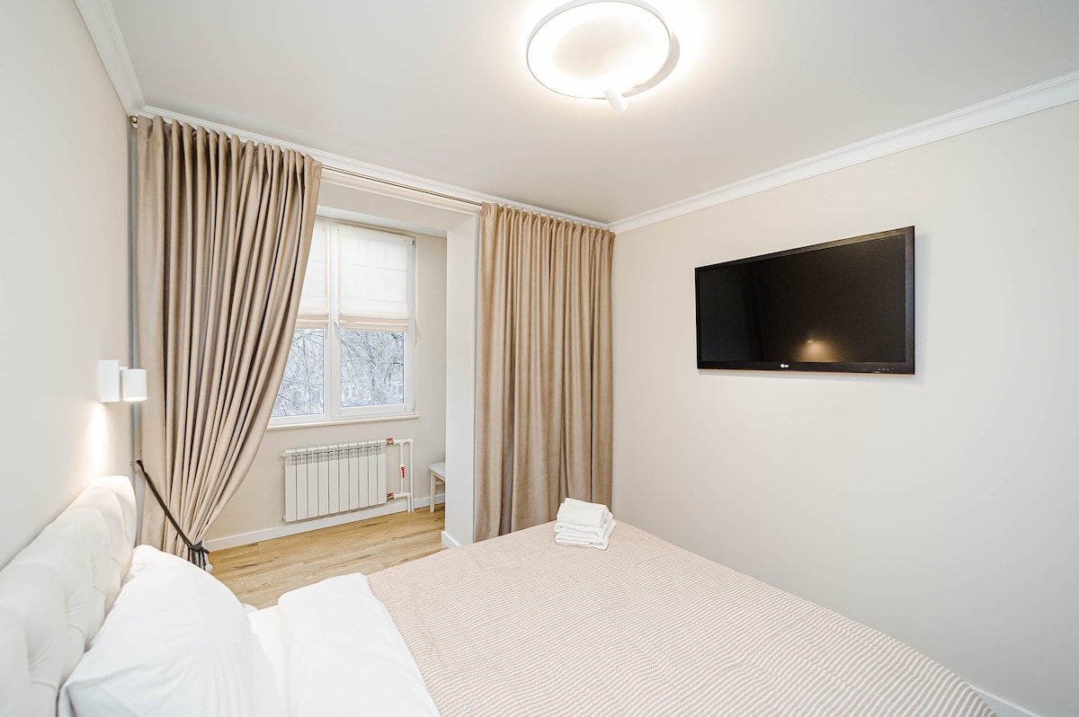 The bedroom features a spacious bed with light-colored bedding. Large curtains frame a window that allows natural light to fill the room. A flat-screen TV is mounted on the wall opposite the bed, with a minimalist style enhancing the overall decor.