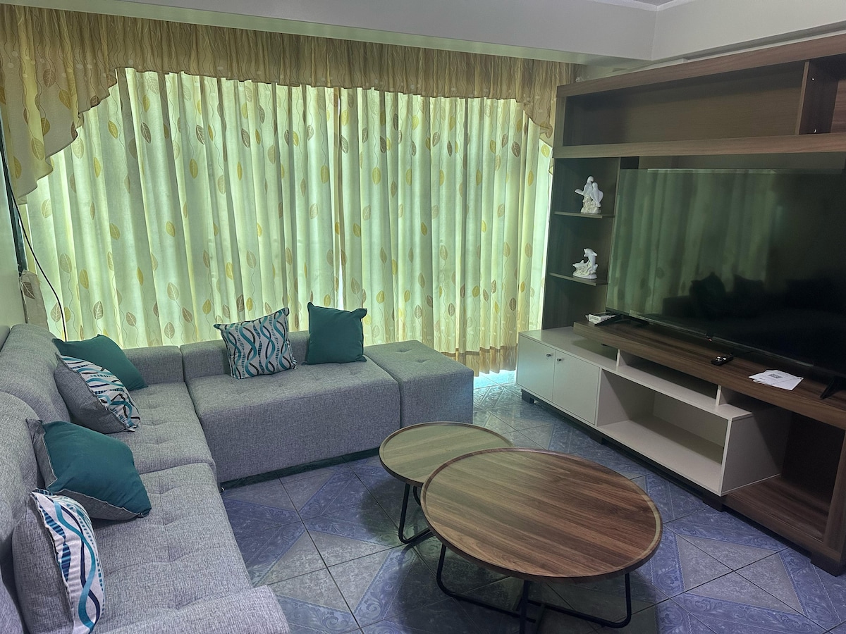 A comfortable living area features a spacious gray sectional sofa adorned with teal and patterned cushions. Two round wooden coffee tables sit on a tiled floor, while large, yellow sheer curtains allow natural light to fill the room and soften the space.