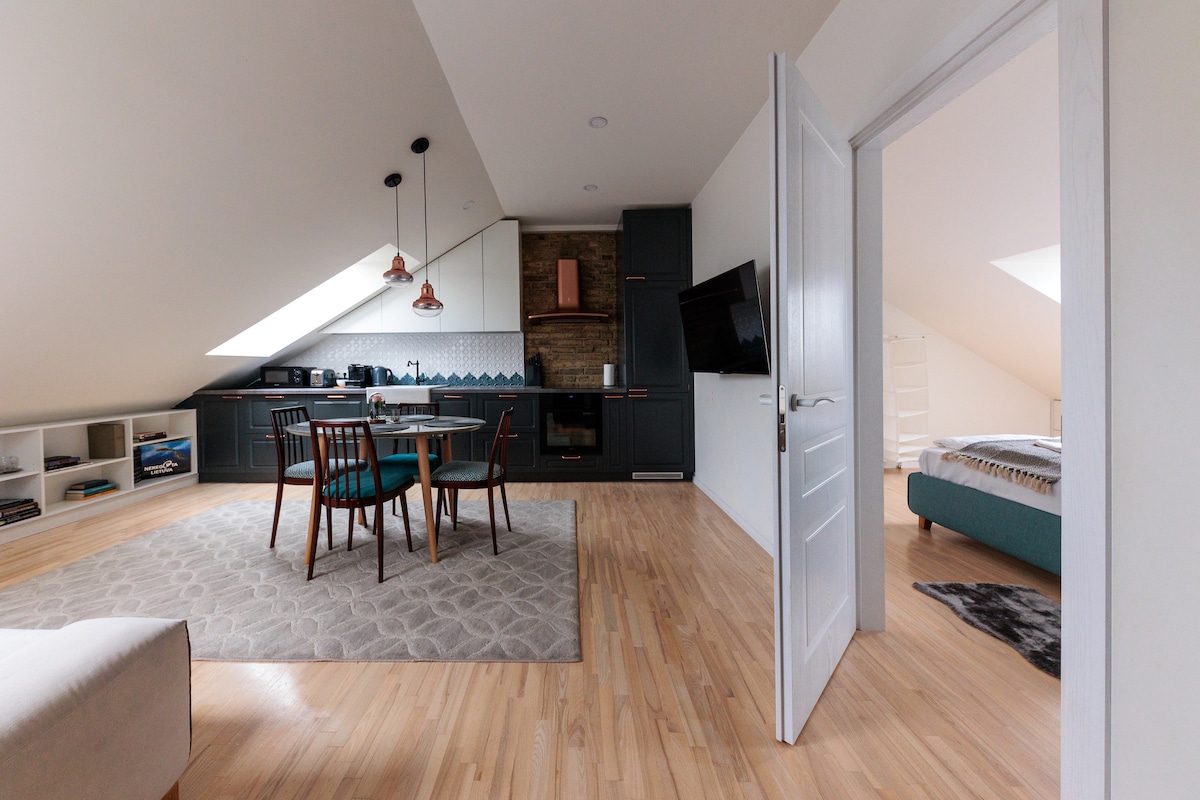 A bright living area is depicted with a light hardwood floor, featuring a dining table surrounded by four chairs. A fully equipped kitchen is visible, complemented by modern appliances and a cozy seating area. Natural light streams in through the sloped ceiling windows.