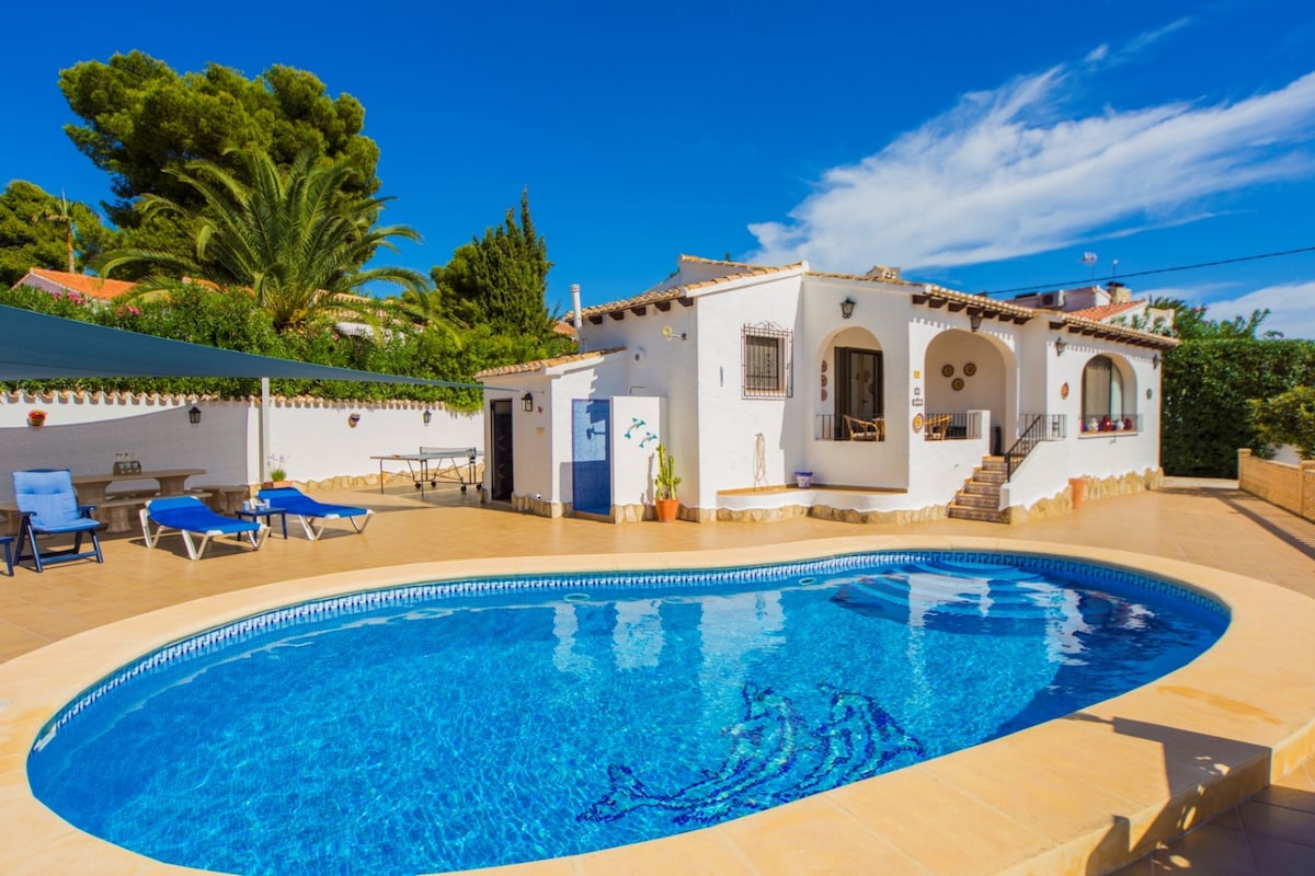 A modernized villa with a heated pool is set in a spacious outdoor area. Sunbeds are positioned near the pool, surrounded by greenery. The villa's exterior features white walls and arched doorways, with a large terrace under a blue sky filled with scattered clouds.