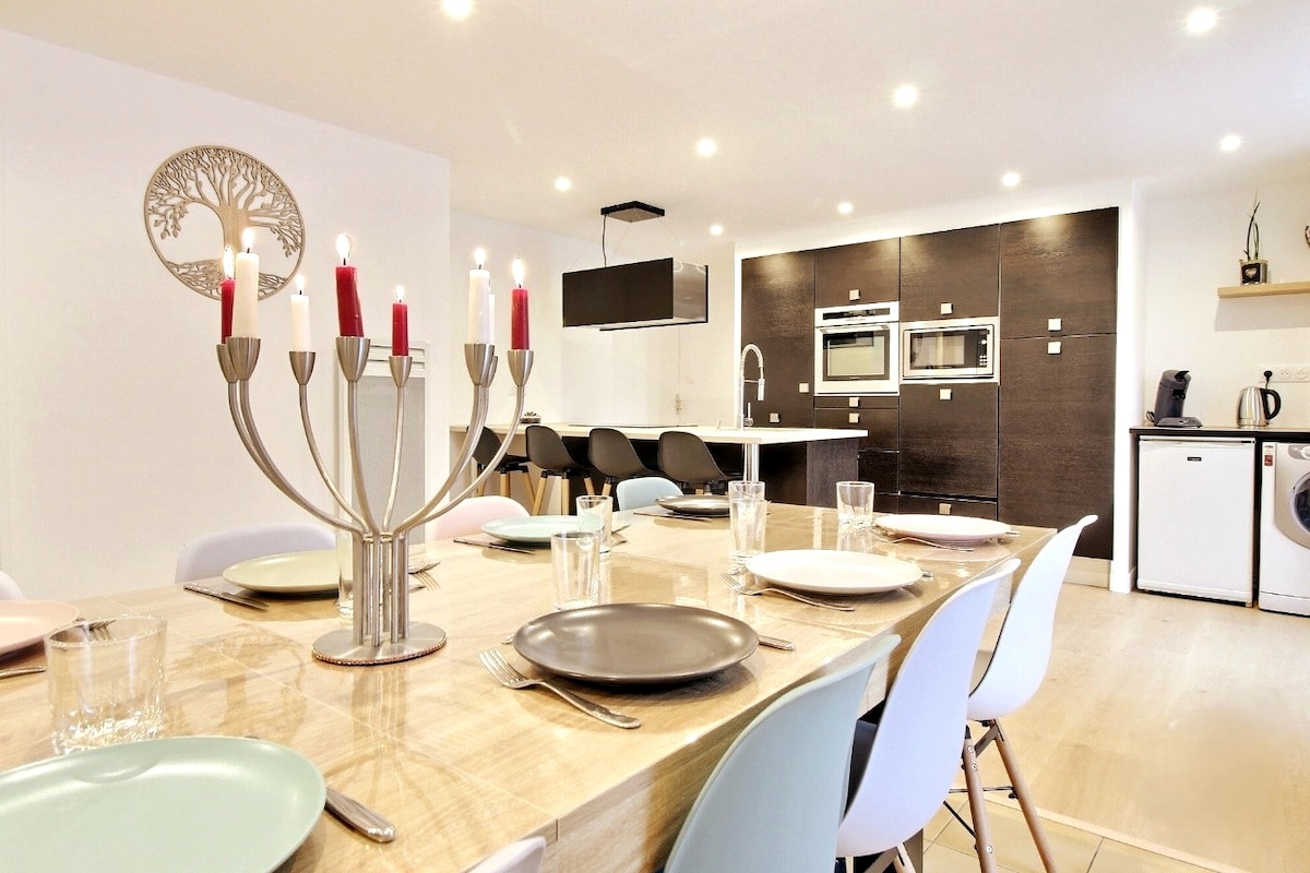 An open-plan kitchen and dining area features a modern design with a large wooden table set for six. Elegant tableware, including plates and glassware, is arranged around a stylish centerpiece candle holder. The kitchen displays sleek cabinetry and built-in appliances, complemented by a clean aesthetic.