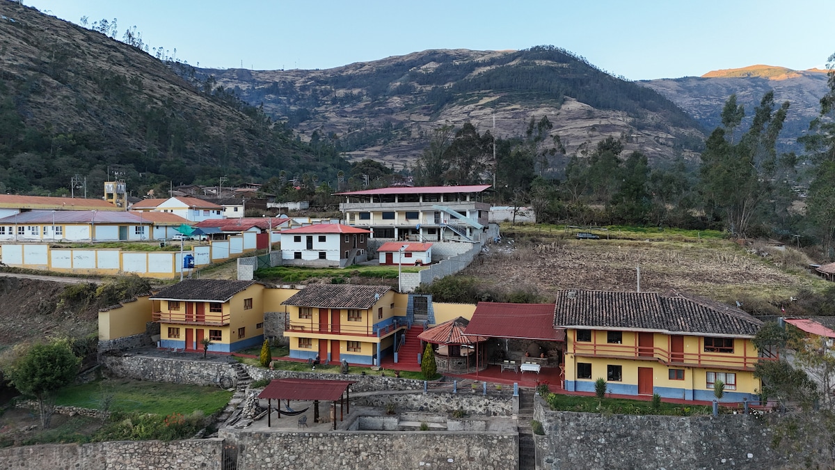 A collection of rustic buildings with terracotta roofs is nestled against a backdrop of rolling hills and mountains. Surrounding lush greenery and cultivated fields can be seen, creating a serene rural landscape. Open outdoor spaces are present for relaxation and enjoyment.