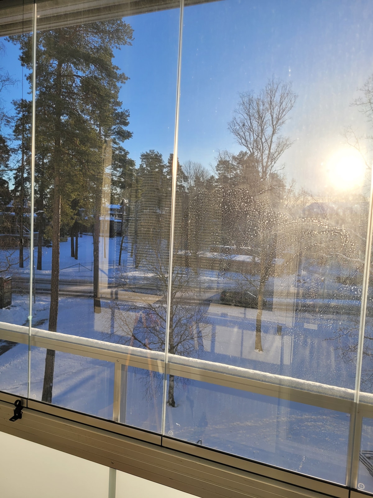 A scenic view is captured through large glass windows, revealing a serene winter landscape. Snow blankets the ground, with tall trees lining the background. The sun casts a warm glow, reflecting off the glass, enhancing the tranquil atmosphere of the outdoor setting.