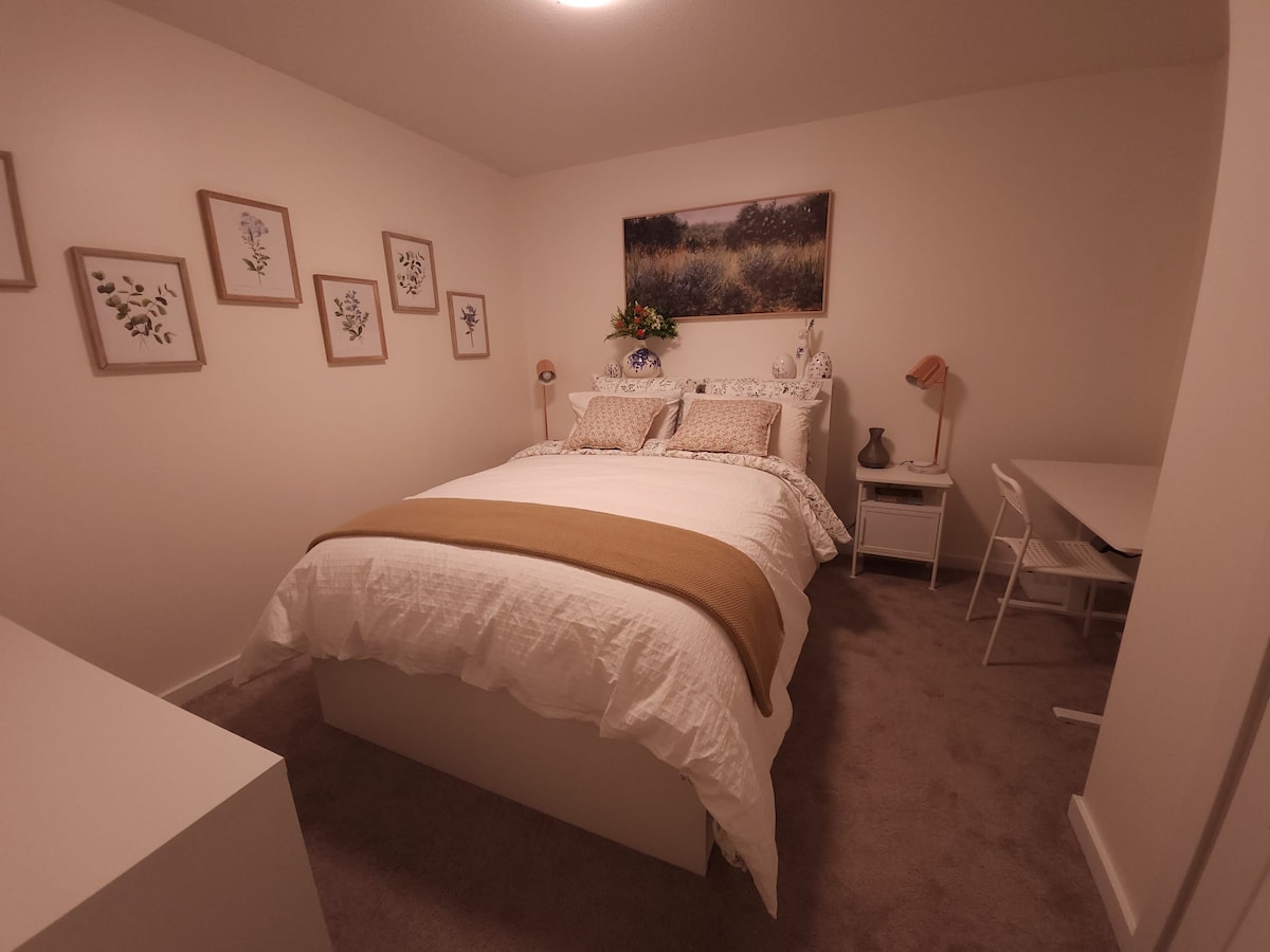 A cozy bedroom features a spacious bed adorned with light bedding and a decorative throw. Framed botanical prints are arranged on the white walls. A small desk and chair are positioned nearby, while a lamp and a vase of fresh flowers add a touch of elegance.