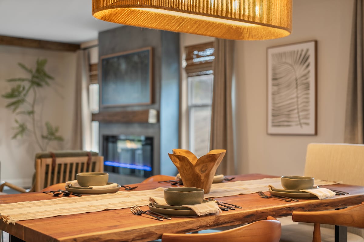 A dining area is showcased with a wooden table set for four, featuring green bowls and cutlery. A decorative centerpiece crafted from wood adds a natural touch. The backdrop includes a cozy fireplace and abstract wall art, enhancing the warm ambiance of the space.
