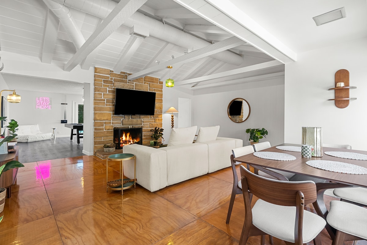 An open-plan living area showcases a light-colored sofa positioned in front of a flat-screen TV mounted above a stone fireplace. A round dining table with dark wooden chairs is visible, and greenery is integrated throughout the space, enhancing a lively yet comfortable setting.