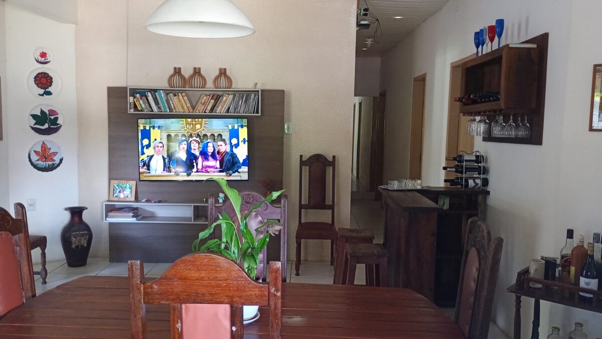 An open dining area features a wooden dining table surrounded by matching chairs. A large television is mounted on the wall, with shelves displaying decorative items and books. A bar unit stands in the corner, displaying various drinks, while a plant adds a touch of greenery.